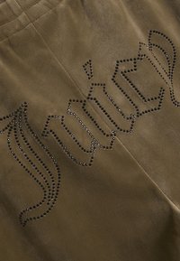 Brązowa tkanina zamszowa z dekoracyjnym wzorem obramowanym małymi czarnymi ćwiekami. Wzór obejmuje zakrzywione linie i wyraźne szczegóły literowe.