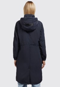 Giacca lunga blu navy con cappuccio, polsini elasticizzati e vita con cordino. Presenta una patch del logo sul retro e una cucitura a spacco sull'orlo.