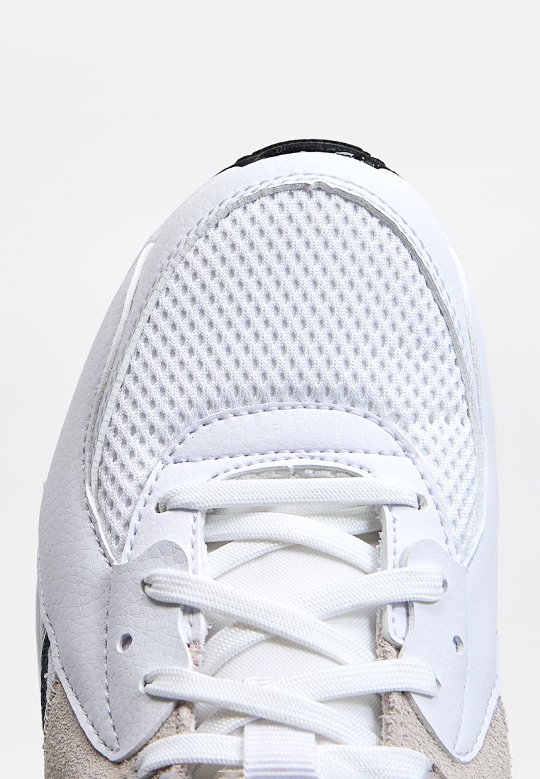 Zapatilla deportiva blanca con textil de malla y cuero sintético, con una puntera texturizada, cordones planos y un acento de ante gris.