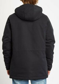 Schwarze Kapuzenjacke mit glatter Textur, geradem Schnitt und elastischen Bündchen. Hat minimale Nähte und wird über hellblauen Jeans getragen.