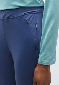 Ναυτικές κολάν με λεία υφή και πλαϊνή τσέπη με φερμουάρ, σε συνδυασμό με ένα ανοιχτό τιρκουάζ μακρυμάνικο τοπ με διακριτική υφή.