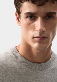 Jasnoszara, dziana sweter z okrągłym dekoltem, o miękkiej teksturze i subtelnych różnicach w grubości, noszony przez męskiego modela.