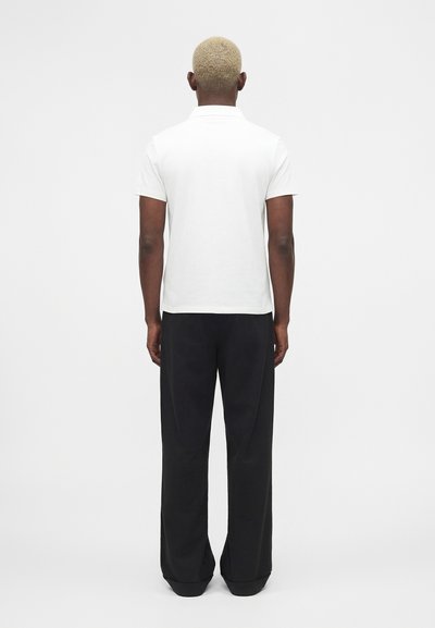 Personne aux cheveux blonds courts vue de dos, portant une chemise blanche à manches courtes et un pantalon noir ample sur un fond uni.