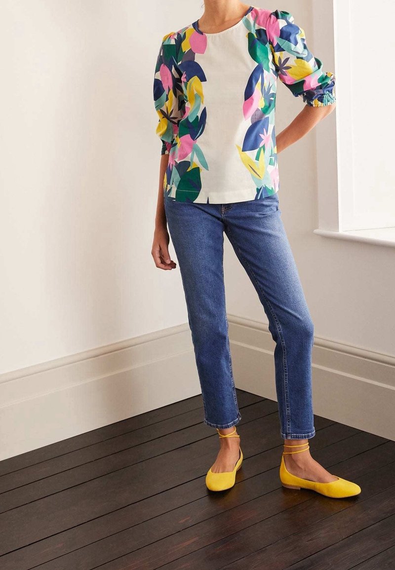 Person standing indoors wearing a floral puff-sleeve top, blue jeans, and yellow ballet flats with ankle straps on dark wooden floor.