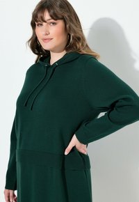 Vestito a maglione verde con cappuccio, con polsini e orlo a costine, caratterizzato da una tasca frontale, realizzato in un morbido materiale a maglia.