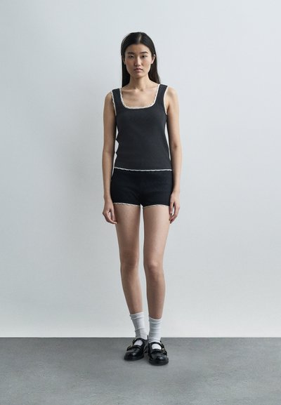 Jeune femme debout contre un mur uni, portant un débardeur noir et un short avec des bordures blanches, des chaussettes blanches et des mocassins noirs.