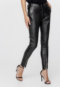 Czarne skórzane spodnie o dopasowanym kroju, z przednimi kieszeniami, detalami szwów oraz zamkami błyskawicznymi przy kostkach. Noszone z czarnymi sandałami na obcasie.