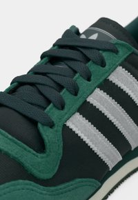 Green and black athletic shoe with a textured mesh upper, three white stripes, thick laces, and a durable rubber sole.