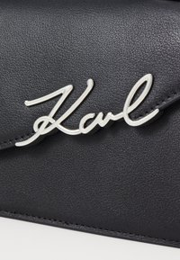 Bolso de cuero sintético negro con un logotipo plateado "Karl" en escritura cursiva. Acabado texturizado con costuras visibles.