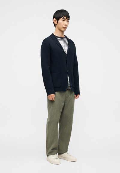 Homme debout portant un blazer bleu marine, une chemise rayée noir et blanc, un pantalon vert olive et des baskets blanches sur un fond blanc uni.