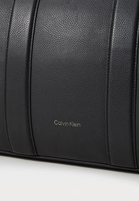 Čierna kožená kabelka s textúrovaným povrchom, s vertikálnymi líniami a zlatým logom "Calvin Klein". Hladké kovové akcenty.