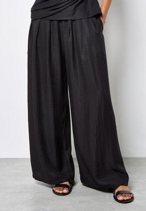 Osoba nosząca luźne, czarne spodnie o szerokich nogawkach oraz czarne, płaskie sandały, stojąca na szarej podłodze na tle białej ściany.