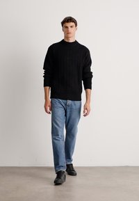 Man walking toward camera wearing black ribbed sweater, light blue jeans, and black sneakers against plain light background.