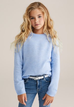 Blonde girl with loose wavy hair wearing a light blue sweater, blue jeans, and a white belt against a plain beige background.