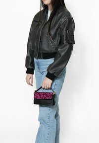 Giacca bomber nera in pelle con polsini a costine, indossata con jeans a zampa d'elefante blu chiaro. Tiene in mano una borsa piccola con un flap a stampa leopardo rosa.