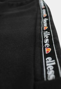 Gros plan sur un tissu noir avec une bande verticale du logo ellesse comportant un texte blanc et un motif de demi-cercle orange-rouge.