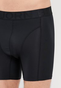 Κοντινή λήψη ενός ατόμου που φορά μαύρες στενές boxer με ορατέ�ς ραφές και φαρδιά ζώνη με ανάγλυφο λογότυπο.