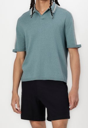 Mężczyzna w tealowym, krótkim polo z dzianiny w białe paski na ramionach oraz czarnych szortach do kolan.