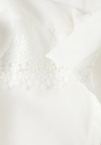 Top de tela blanca con bordado floral detallado en el dobladillo, con textura suave y sutil brillo, creando una apariencia de capas.