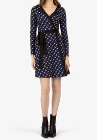 Blaues Wickelkleid mit einem geometrischen Muster in Schwarz und Creme, langen Ärmeln, einem gebundenen Taillenbund und einer glatten Textur. Getragen mit schwarzen Stiefeletten.