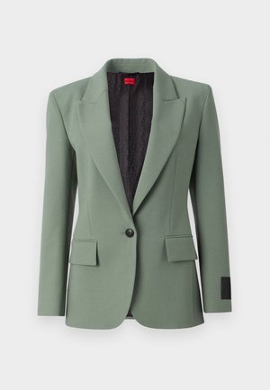 Lys grønn blazer laget av glatt stoff, med oppslag, enkelt knappelukking, to frontlommer og et mønstrete svart fôr.