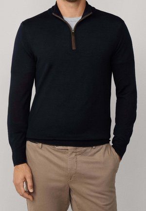 Homme portant un pull noir à fermeture éclair au quart sur une chemise grise avec un pantalon beige, main gauche dans la poche, sur fond uni.