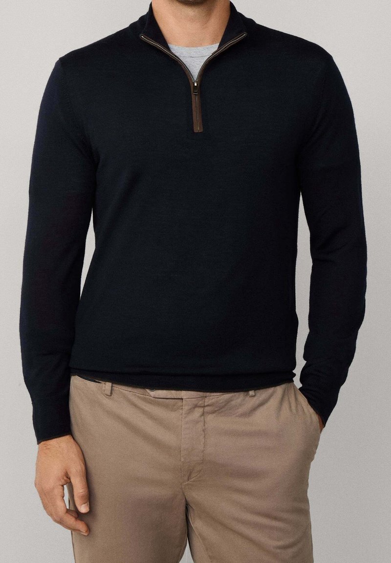 Homme portant un pull noir à fermeture éclair au quart sur une chemise grise avec un pantalon beige, main gauche dans la poche, sur fond uni.