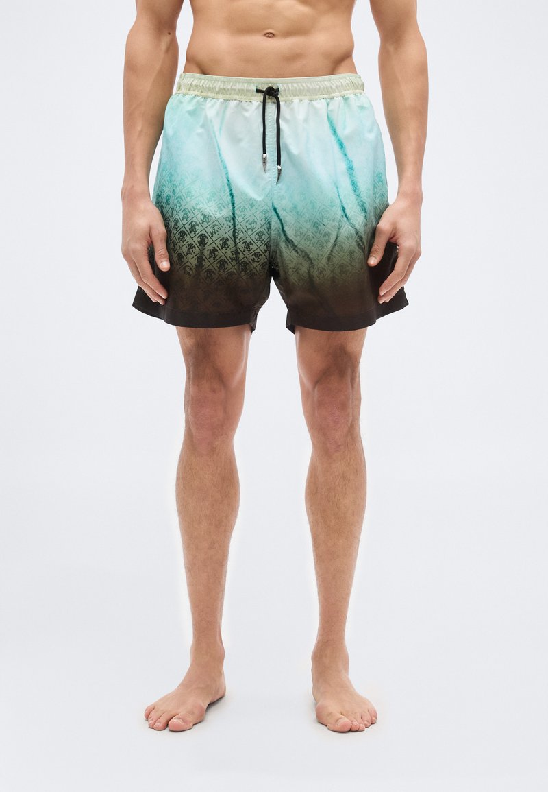 Άνδρας με μαγιό σε απόχρωση βαθμια ίου μπλε-πράσινου και καφέ με μαύρο κορδόνι, όρθιος με γυμνά πόδια μπροστά σε απλό λευκό φόντο.