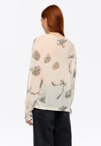 Lekka bluzka z długim rękawem w kolorze beżowym z wzorami róż i kokardek w stylu akwareli. Materiał ma miękką, przejrzystą teksturę i luźny krój.