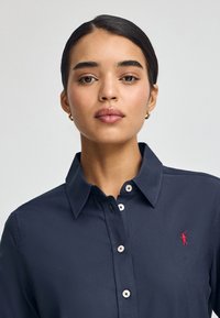 Granatowa koszula zapinana na guziki wykonana z gładkiej bawełny. Posiada wyrazisty kołnierz oraz białe guziki, ozdobiona małym czerwonym logo po lewej stronie klatki piersiowej.