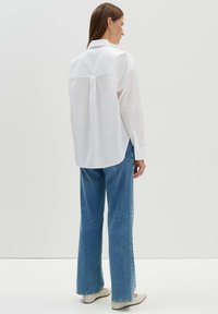 Camicia bianca con colletto a yoke sul retro, abbinata a jeans a gamba larga di colore blu chiaro. La camicia ha maniche lunghe e un orlo curvo.