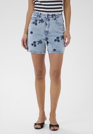 Kvinde iført lyseblå denimshorts med mørke blomsterprint, kombineret med sorte sandaletter med åben tå og en stribet top uden ærmer.