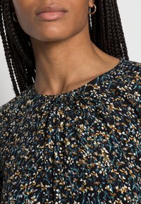 Schwarze Bluse mit gerafftem Ausschnitt, die ein florales Muster in Blau, Gelb und Weiß auf einem dunklen Hintergrund zeigt. Glatte Stoffstruktur.
