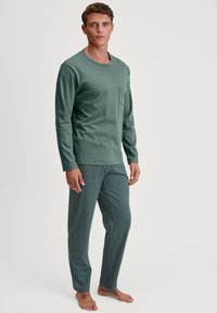 Lange mouwen groene shirt met zak, gecombineerd met gestreepte groene en zwarte pyjamabroek met geometrische vormen. Model is blootvoets.