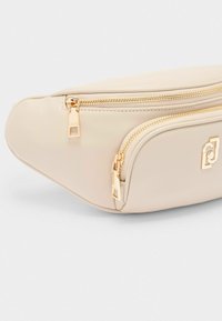 Marsupio in finta pelle beige con due scomparti con cerniera e dettagli in hardware dorato. Forma arrotondata con una texture liscia e opaca.