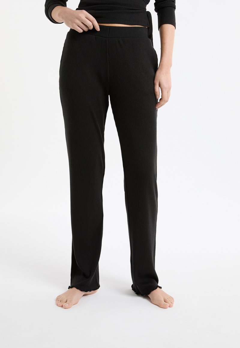 Mujer con pantalones negros holgados de tejido acanalado y una camiseta negra a juego, de pie descalza sobre un fondo blanco.