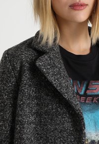 Grijze, gestructureerde wollen jas met een reverskraag, gedragen over een zwart grafisch t-shirt. Focus op de stof en de gestructureerde vorm van de jas.