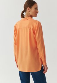 Long-sleeve, button-up shirt in bright orange fabric, featuring a relaxed fit, a curved hem, and visible stitching on the back yoke.