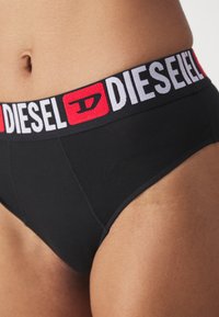 Fekete pamut bugyi fehér és piros elasztikus derékbővítéssel, rajta a "DIESEL" logóval. Simára csiszolt textúrával és testhezálló fazonnal rendelkezik.
