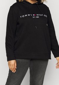 Person wearing black Tommy Hilfiger hoodie with logo and "EST. 1985" text, paired with dark gray jeans, hands relaxed at sides.