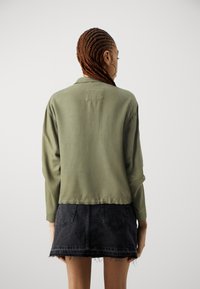 Roheline pikakätine särk, millel on veidi lühem lõige, pehme kangas ja seljaääres detail, koos musta denimsinise seeliku ja friteeritud servaga.