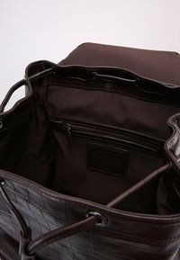 Hnědá taška z umělé kůže s prošívanou texturou, měkkým vnitřkem a vnitřní kapsou na zip. Dvojité uši pro snadné nošení.