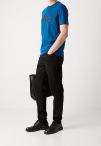 Camiseta de algodón azul con el logo en negro "BOSS", combinada con pantalones negros y zapatos de cuero negros, sosteniendo un maletín negro.