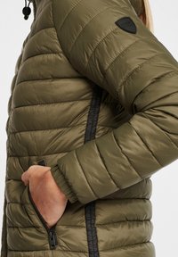 Veste matelassée vert olive avec coutures horizontales, poche latérale zippée et accents noirs sur la fermeture éclair et la pièce d'épaule.