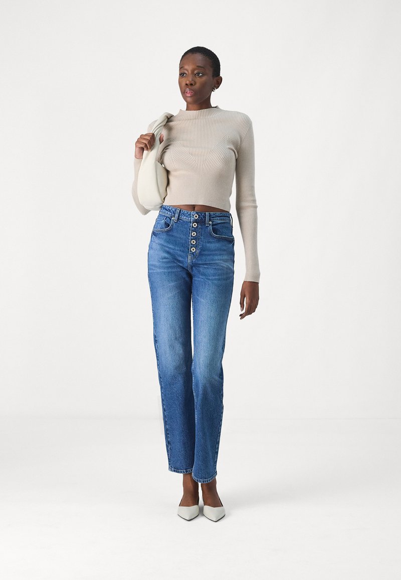 Beige gerippter Crop-Pullover, hochtaillierte blaue Jeans mit Knopfleiste und weiße spitze Schuhe. Modell hält eine leichte Handtasche.