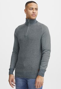 Gray half-zip sweater with a textured pattern, ribbed cuffs, and a contrasting brown label at the hem. Worn with light blue jeans.