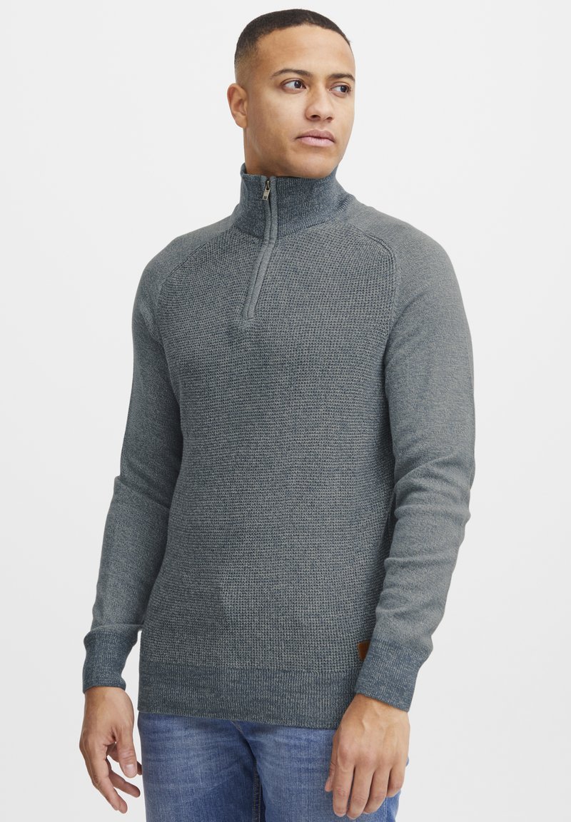 Gray half-zip sweater with a textured pattern, ribbed cuffs, and a contrasting brown label at the hem. Worn with light blue jeans.