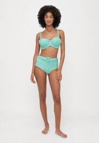 Τετράγωνο μπικίνι σε τουρκουάζ απόχρωση με γυαλιστερή υφή, που διαθέτει μια δομημένη μπλούζα με φαρδιές τιράντες και ψηλόμεσα σλιπ με ζώνη και διακοσμητικό κουμπί σε σχήμα κύκλου.