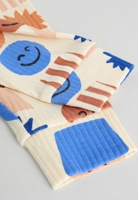 Sokszínű bordázott zoknik absztrakt formákkal és mosolygó arcokkal kék, narancs és barna színekben, világos krémszínű háttéren.