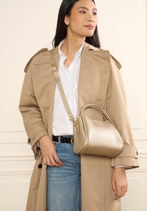 Femme portant un trench-coat beige et une chemise blanche tient un sac à main doré métallique avec bandoulière, debout contre un mur crème.